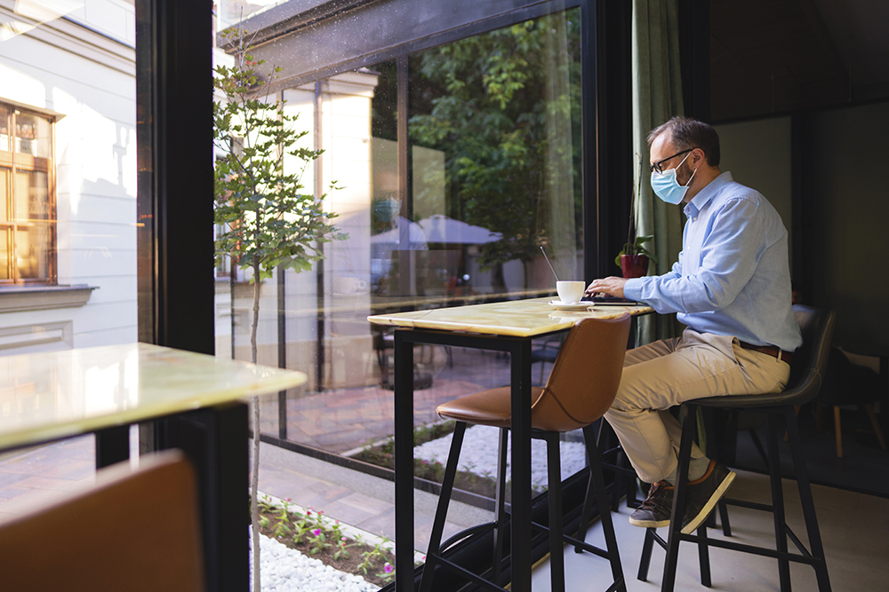 一个戴着防护口罩的成熟白人商人坐在咖啡馆里，用他的笔记本电脑工作。